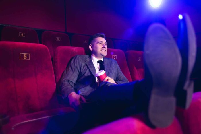 młody, przystojny mężczyzna siedzący samotnie w kinie, w garniturze służbowym, z nogami na przednim siedzeniu